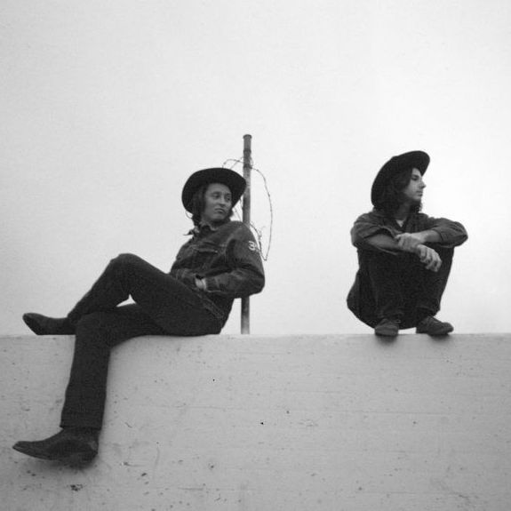 Two members of Mapache photographed atop a white wall, one seated, one crouching.