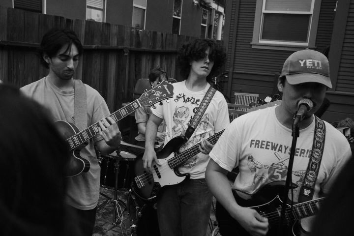 Three young male musicians in t-shirts, wearing guitars (one bass, two six strings), playing music outside in what appears to be a back yard, with a corner of a house and a wooden fence visible.