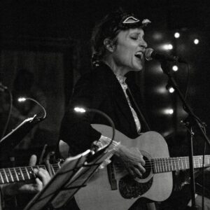 Image of musician Deborah Crooks playing a guitar and singing in black in
