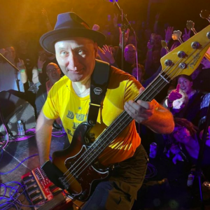 Jah Wobble playing bass live on stage with crowd in the background.