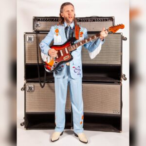 Jimmy Touzel in a blue suit holding an electric guitar in front of multiple amps