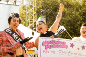 The winner of a drag contest smiling being presented with a large check from the former winner