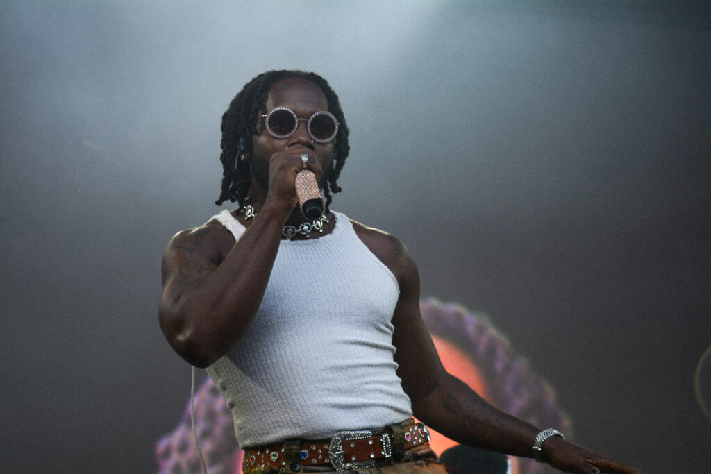 Man in a white tank top and dark sunglasses singing into a microphone on stage