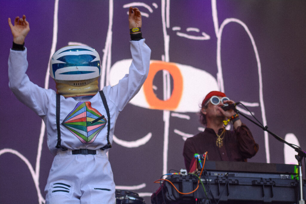 A band performing on stage with one member in an astronaut suit and one standing at a keyboard singing into a microphone