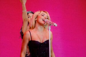 Performer singing into a sequin covered microphone with a dancer in the background and a bright pink backdrop