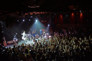 Rock band performing on stage with drums, bass, guitar, and singer in front of a clapping crowd