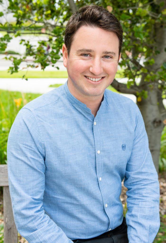 Man wearing a blue button down shift standing outside near a tree.