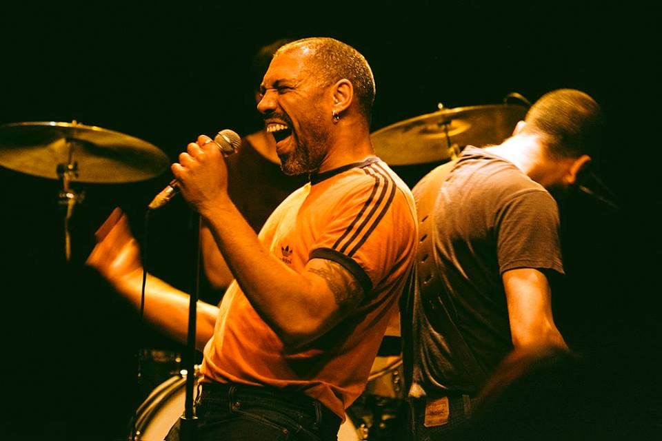 vocalist, guitarist, and drummer all performing in an orange glow