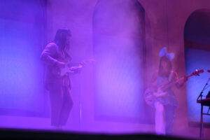 Two guitarists, one man in a tan suit and one woman in a blue dress and large bow on stage in fog