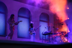 Band performing on stage, from left to right man playing guitar, woman playing guitar, an unattended keyboard, and man playing drums