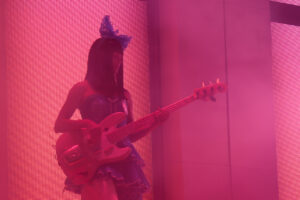 Guitarist performing on stage wearing a blue dress and large bow in her hair in pink stage lighting