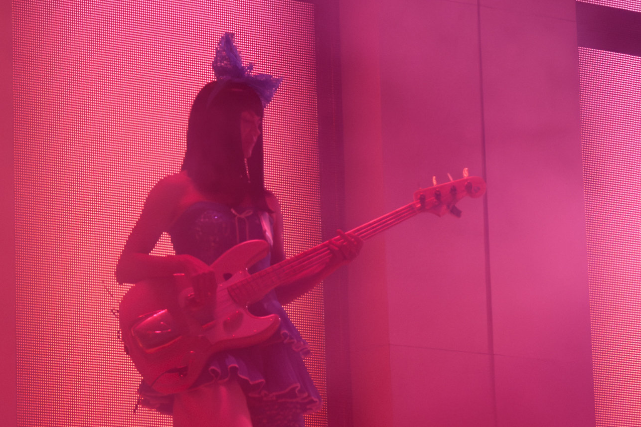 Guitarist performing on stage wearing a blue dress and large bow in her hair in pink stage lighting