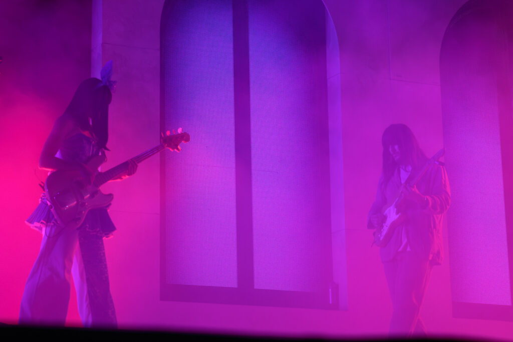 Two guitarists, one man in a tan suit and one woman in a blue dress and large bow on stage in fog