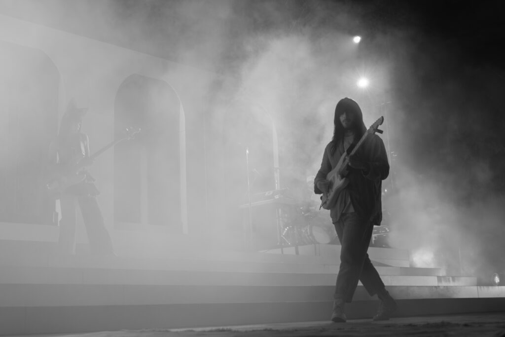 Two guitarists, one man in a tan suit and one woman in a blue dress and large bow on stage in fog in black and white