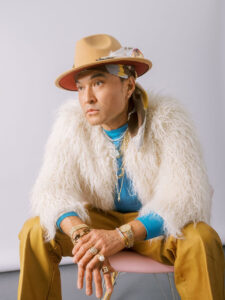 A man sitting with arms crossed, wearing a hat, blue shirt, white fur, and brown pants.