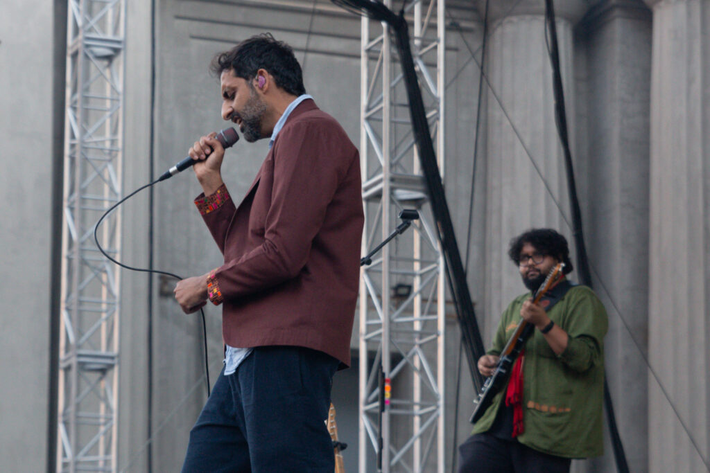 Man singing into a microphone in front of the band's guitarist on stage during the day