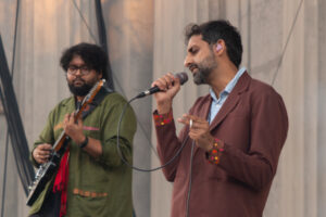 Man singing into a microphone, holding a cigarette in the other hand, in front of the band's guitarist on stage during the day