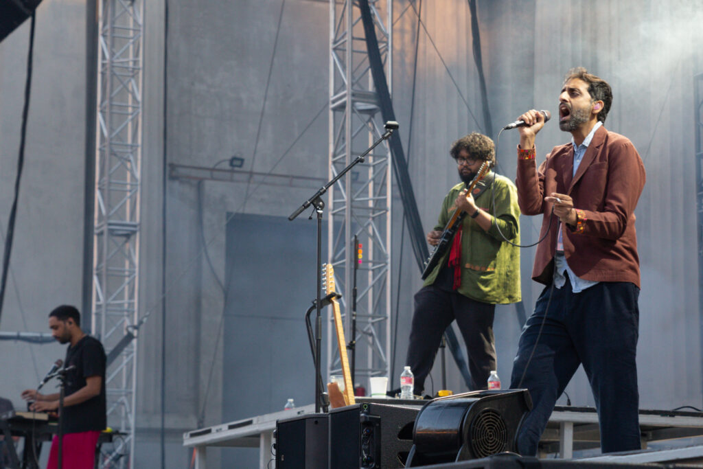 Man singing into a microphone in front of the band's guitarist and keyboardist on stage during the day