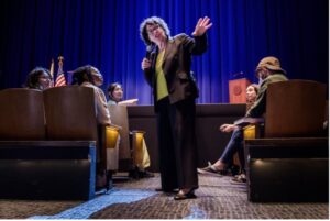 Supreme Court Justice Sonia Sotomayor standing at the floor level of an auditorium among students who sit looking on. She is speaking into a microphone with the other hand raised.