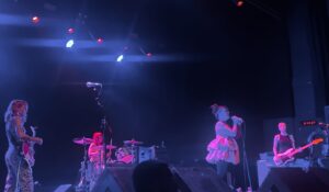 The punk band Bikini Kill performing on stage featuring the drummer, two guitarists, and vocalist within view