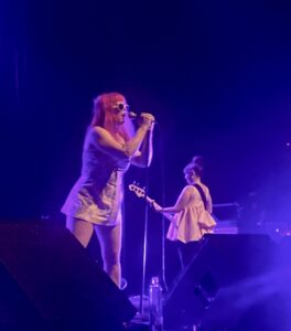 Singer leaning into a microphone with a guitarist in the background while performing on stage