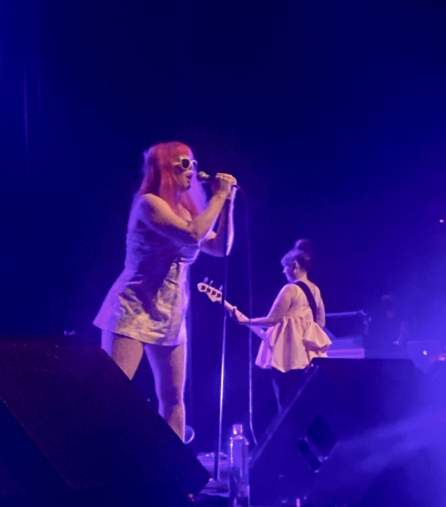 Singer leaning into a microphone with a guitarist in the background while performing on stage