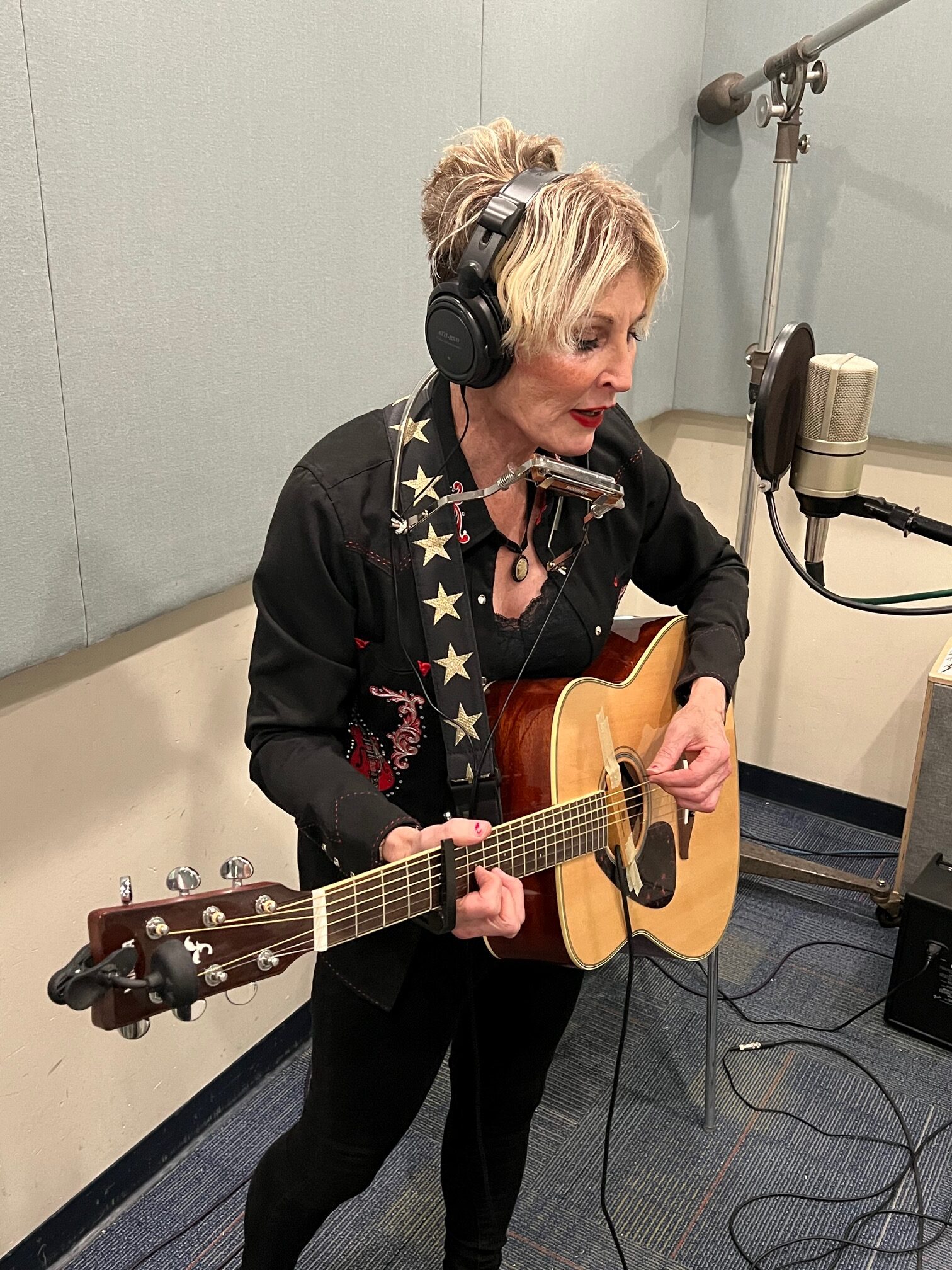 Woman performing in a studio singing into a microphone and playing guitar with a harmonica around her neck.