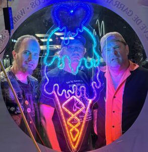 Three men standing on the other side of an ice cream shop window through a neon ice cream cone sign.