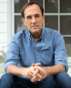 Man, wearing a blue shirt and jeans, sitting in front of a window with hands folded.