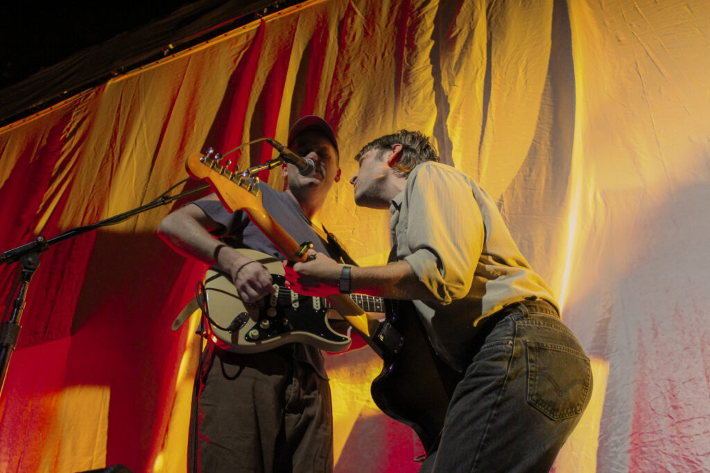 Two musicians playing electric guitar on stage both singing into the same microphone.