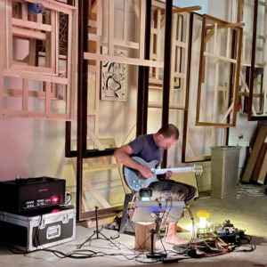 John Davis playing guitar in a room with frames behind him.