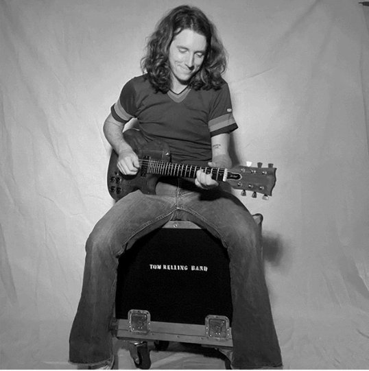 Black and white press photo of musician Tom Relling sitting on an amp playing a guitar