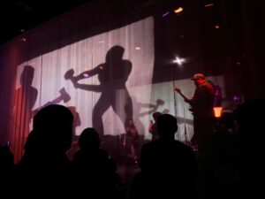 A rock band playing on stage as two figures pound hammers from behind a curtain on stage