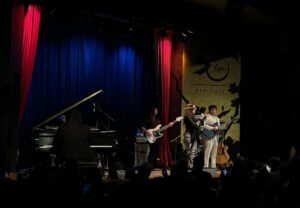 A vocalist performing on stage alongside two guitarist and a pianist, taken from the back of a seated crowd.