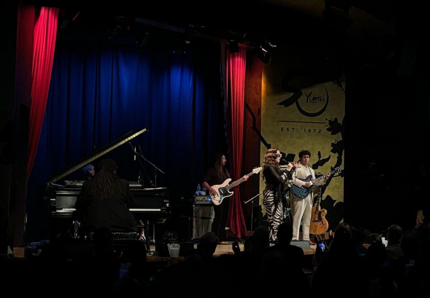 A vocalist performing on stage alongside two guitarist and a pianist, taken from the back of a seated crowd.