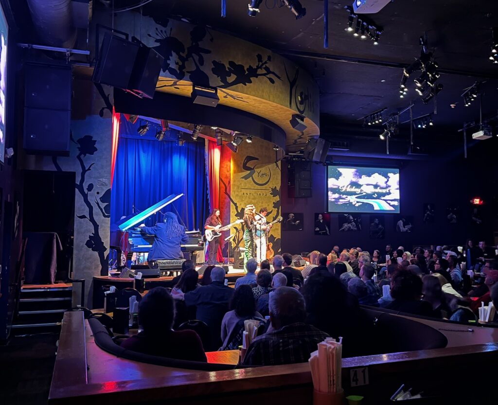 A vocalist performing on stage alongside two guitarist and a pianist, taken from the back of a seated crowd.