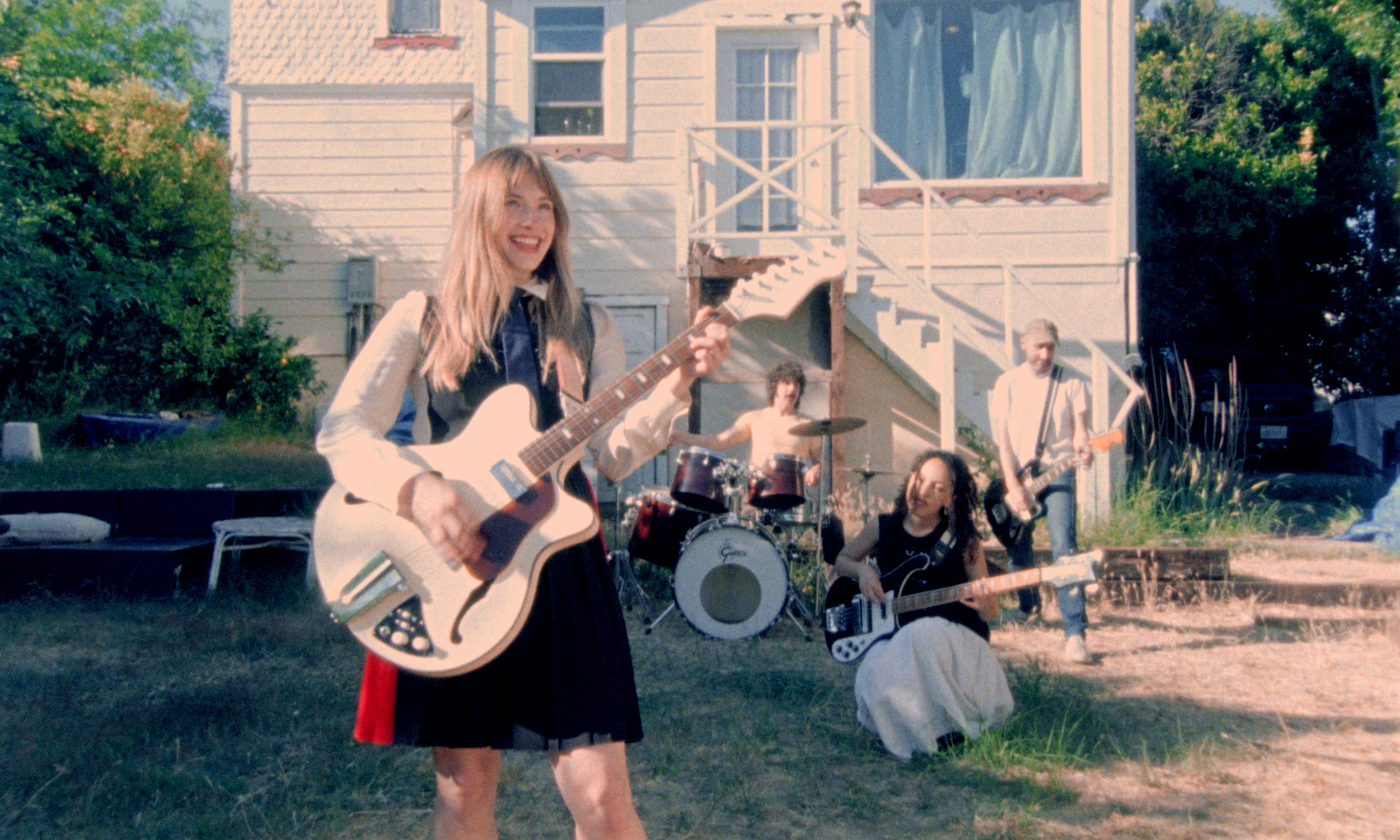 Musician Kate Bollinger playing guitar in front of her band outside beyond a house