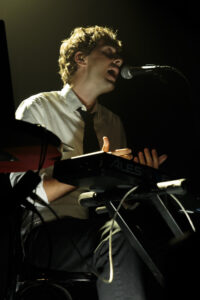 A man sitting over a synthesizer, hands raised chest high, while signing