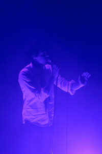 A man in a white shirt and tie singing in to a microphone bathed in blue light