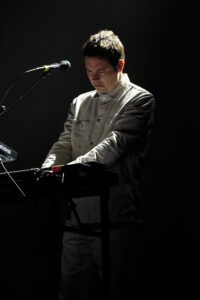 A man, dressed in all grey, playing an electronic instrument on stage