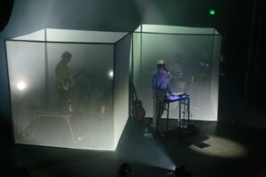 A band on stage with white light over head, one member exposed with two behind encased in a square screen obscuring their view