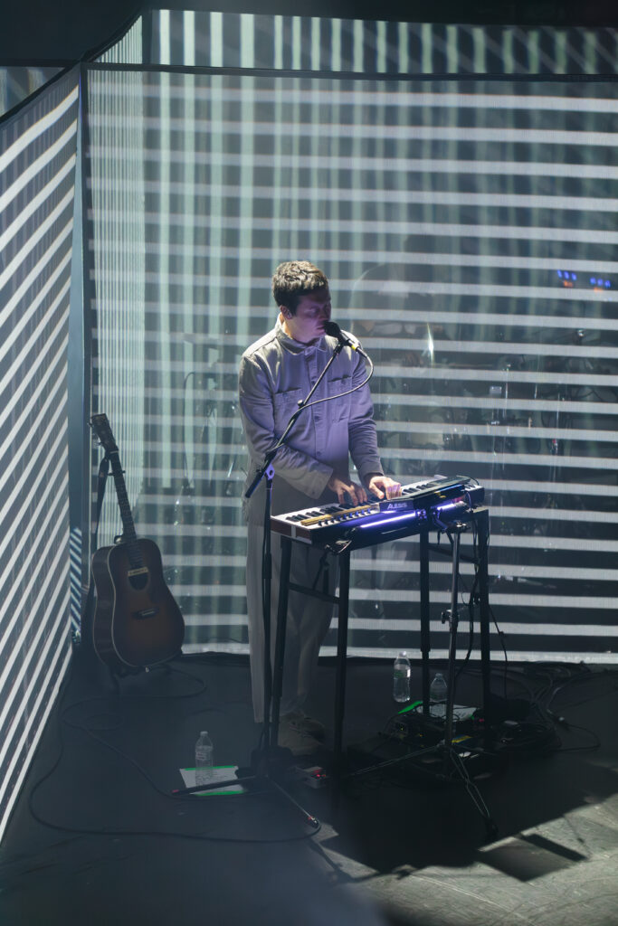 A man, dressed in all grey, playing an electronic instrument on stage next to an acoustic guitar