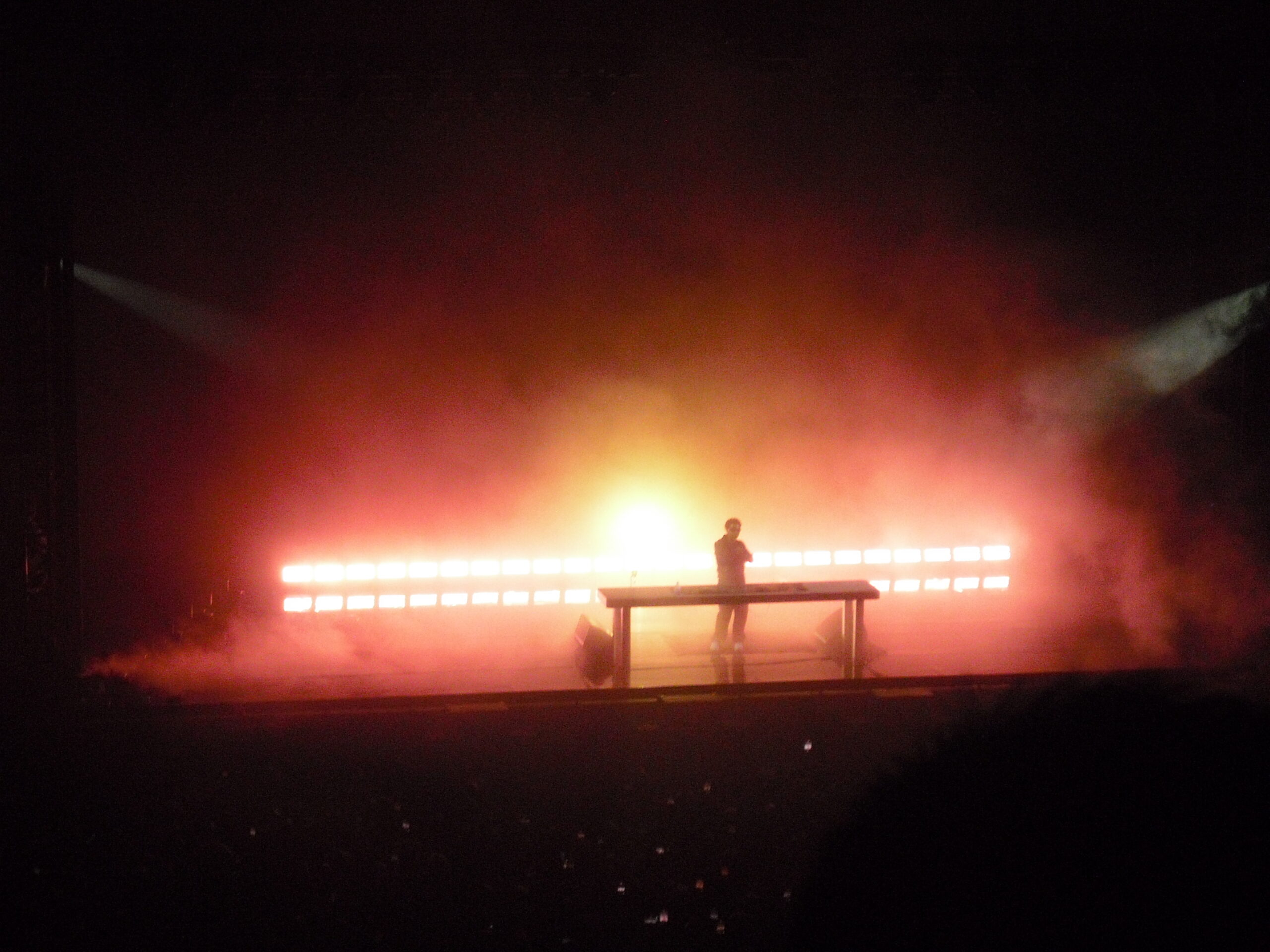 Kaytranada standing arms crossed behind his electronic equipment on stage with an orange glow