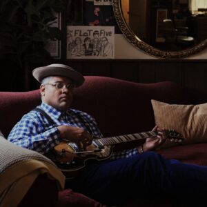 Dom Flemons sitting on a couch with a guitar.