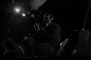 Black and white photo of Robin Pecknold sitting, singing, and playing guitar on stage