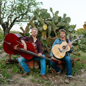 Two men sitting in front of cactus outside holding guitars